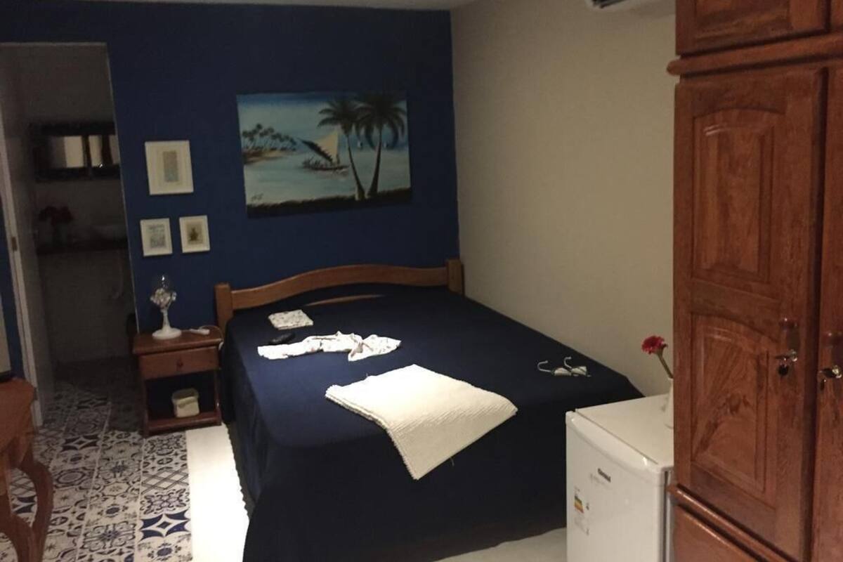 Un dormitorio con cama, mesita de noche, cómoda, un cuadro y un electrodoméstico blanco.