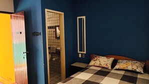 A bedroom with a checkered bedspread, a wooden headboard, a mirror on the wall, and a door leading to a bathroom. - Pousada 100 Passos (Tamandaré)