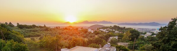 Paisagem costeira com um pôr do sol, uma montanha e uma cidade ao longe.