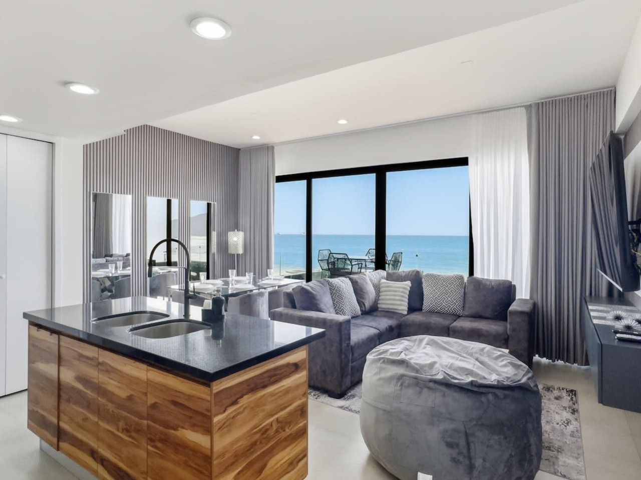 A modern living room with a large window, a sofa, a coffee table, and a kitchen area with a wooden island.