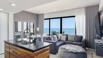 A modern living room with a large window, a sofa, a coffee table, and a kitchen area with a wooden island.