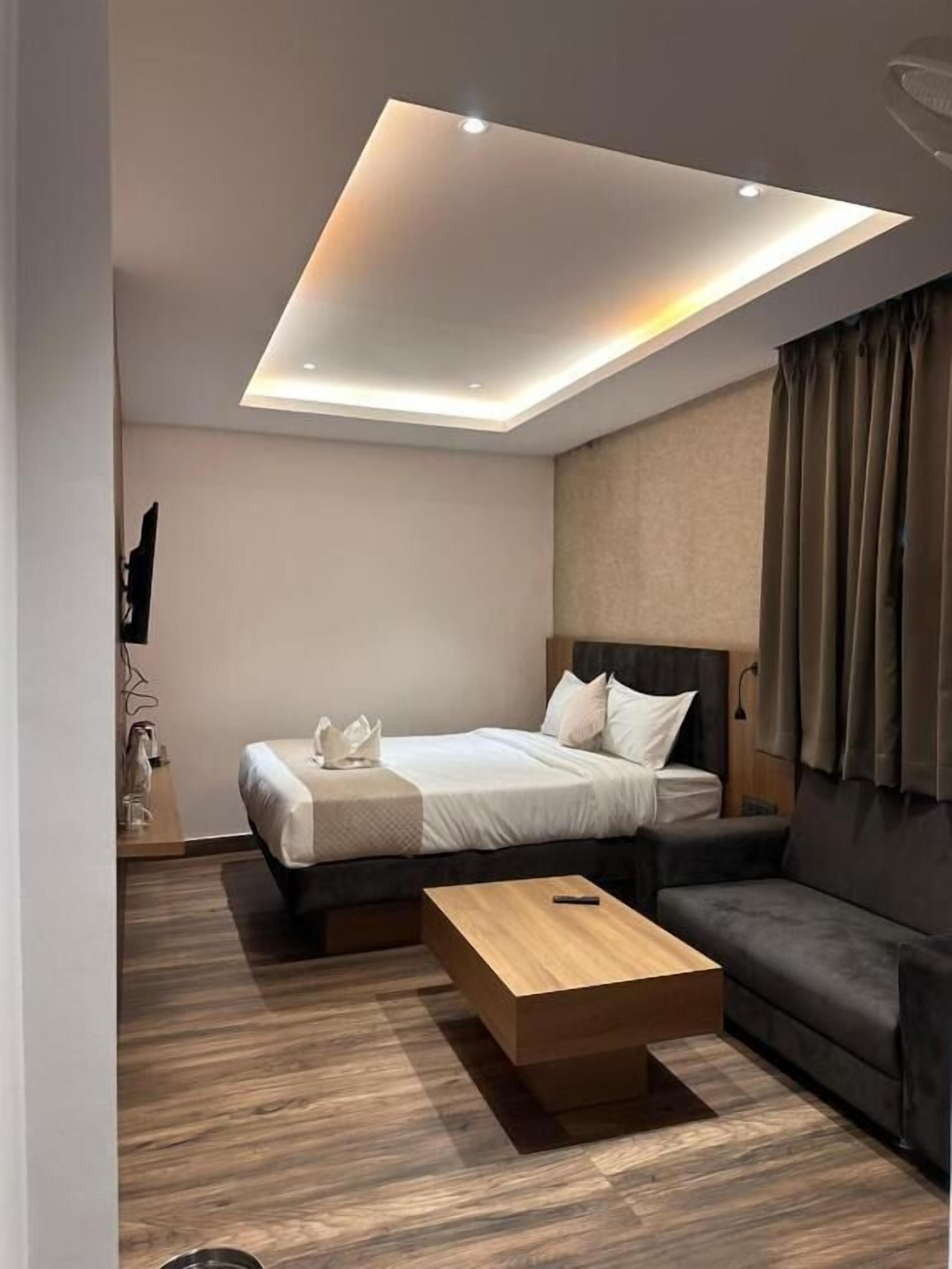 A modern hotel room with a bed, a sofa, a wooden coffee table, and a recessed ceiling light.