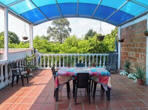 Eine überdachte Terrasse mit Tisch und Stühlen, umgeben von Topfpflanzen und einem weißen Balkon.