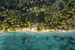 Vista aérea de uma praia com águas turquesas cristalinas, palmeiras e um pequeno conjunto de edifícios.