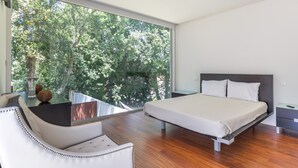 Un dormitorio moderno con un ventanal grande, una cama, una silla y una mesita.