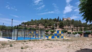 A sandy area with a blue and yellow play structure, a fenced pool, and a building on a hill in the background.