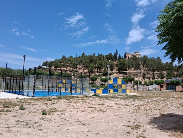 Zona arenosa con una estructura de juego azul y amarilla, piscina cercada y un edificio en la distancia sobre una colina.