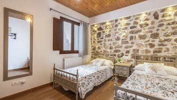 A bedroom with two beds, a stone wall, wooden flooring, a mirror, and a window with curtains.