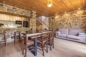 Un interior rústico con pared de piedra, techo de madera, zona de comedor con sillas y mesa, un sofá y una cocina con electrodomésticos.