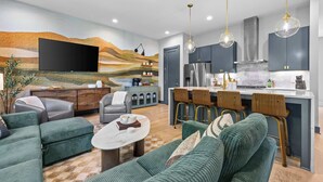 Een moderne woonkamer met een flatscreen televisie, groene banken, een salontafel en een keuken met een koelkast, fornuis en barstoelen.