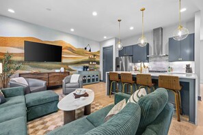 Ein modernes Wohnzimmer mit Flachbildfernseher, grünen Sofas, einem Couchtisch und einer Küchenzeile mit Kühlschrank, Herd und Barhocker.
