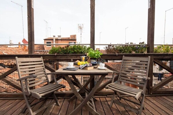 Eine Holzterrasse mit zwei Stühlen, einem Tisch, einem Blumentopf und Blick auf Gebäude im Hintergrund.
