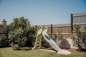 A playground with a slide and climbing structure in a grassy area with a wooden fence and bushes in the background. - House with ocean views, Port de Doëlan, Le Lodge de Keruster, 4 bedrooms (Clohars-Carnoët)