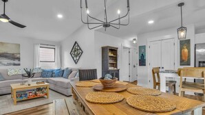 A modern living room with a wooden dining table, a sofa set, and a kitchen area with white cabinets. - The Good Life OKI | Hot Tub & Game Room (Oak Island)