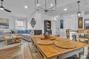 A modern living room with a wooden dining table, a sofa set, and a kitchen area with white cabinets. - The Good Life OKI | Hot Tub & Game Room (Oak Island)