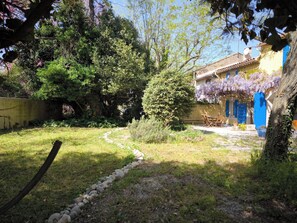 Un jardin avec un chemin de pierres, un banc en bois et un bâtiment aux portes bleues et aux fleurs violettes.