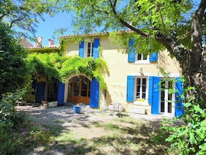 Un bâtiment jaune avec des volets bleus, une entrée abritée et un jardin agrémenté d’une table et de chaises.