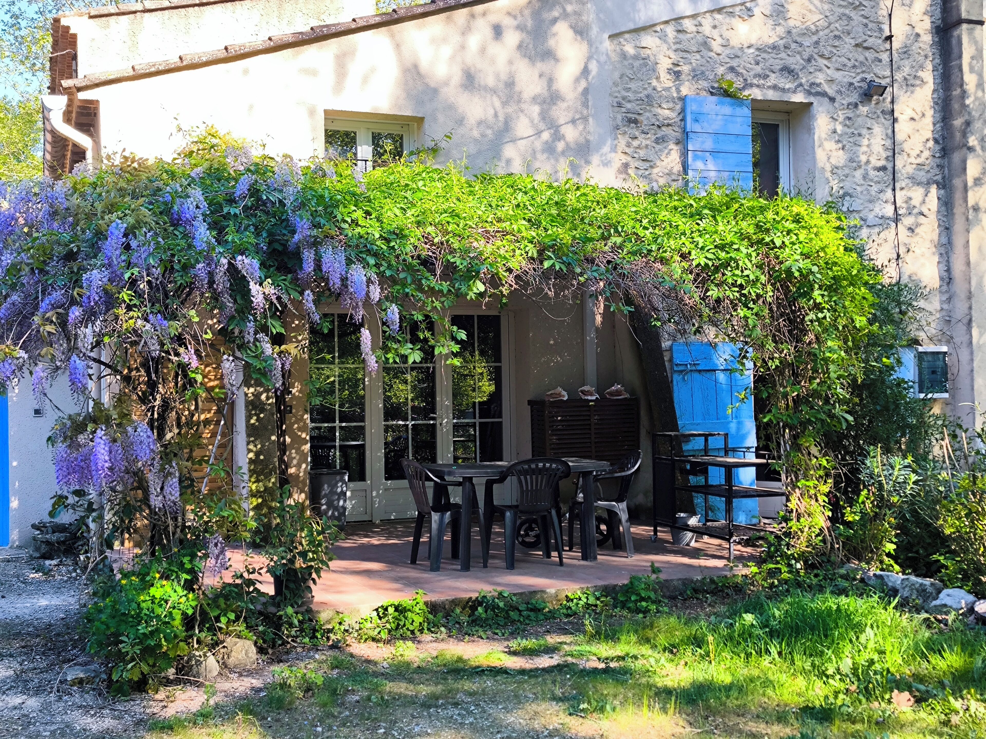 Une terrasse avec une table et des chaises, un espace couvert offrant une vue sur un bâtiment, et un jardin agrémenté de fleurs violettes.