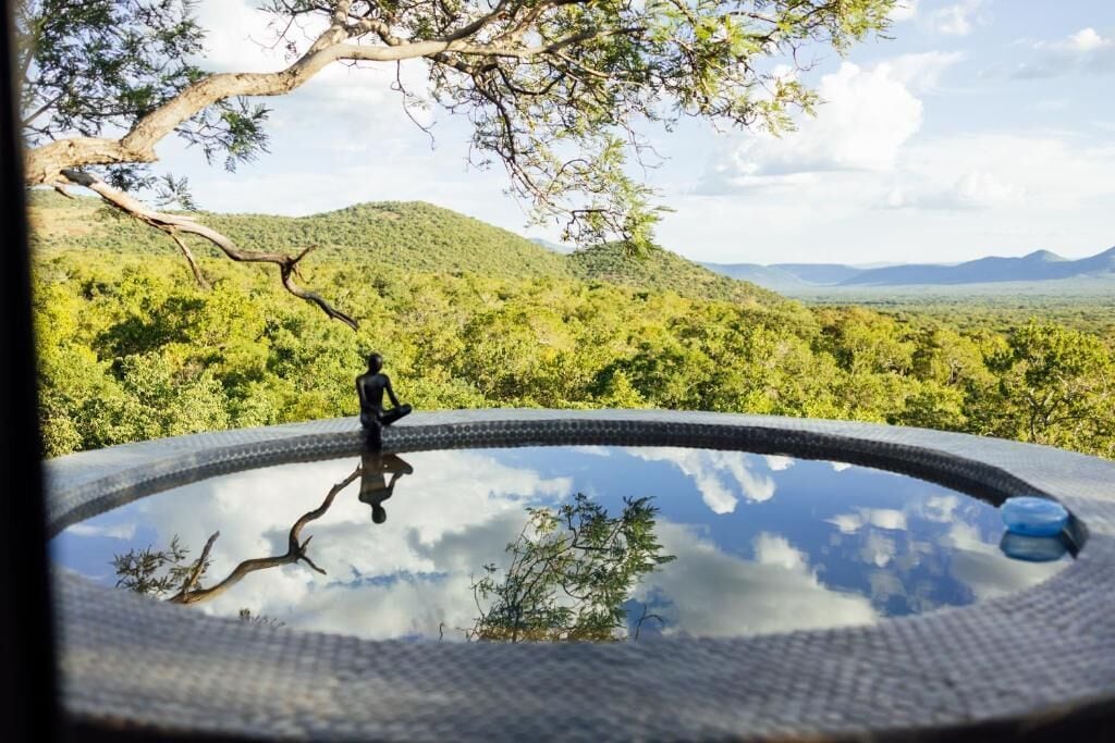 Una piscina circular con una persona meditando, rodeada de abundante vegetación y colinas.