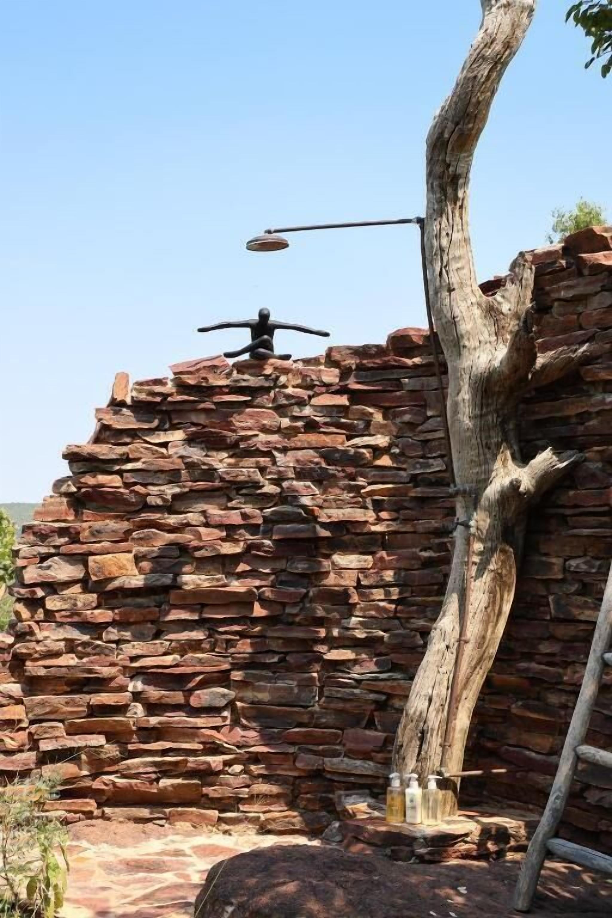 Un mur de pierres rustiques percé par un arbre qui le traverse, un lampadaire en métal et une échelle appuyée contre le mur.