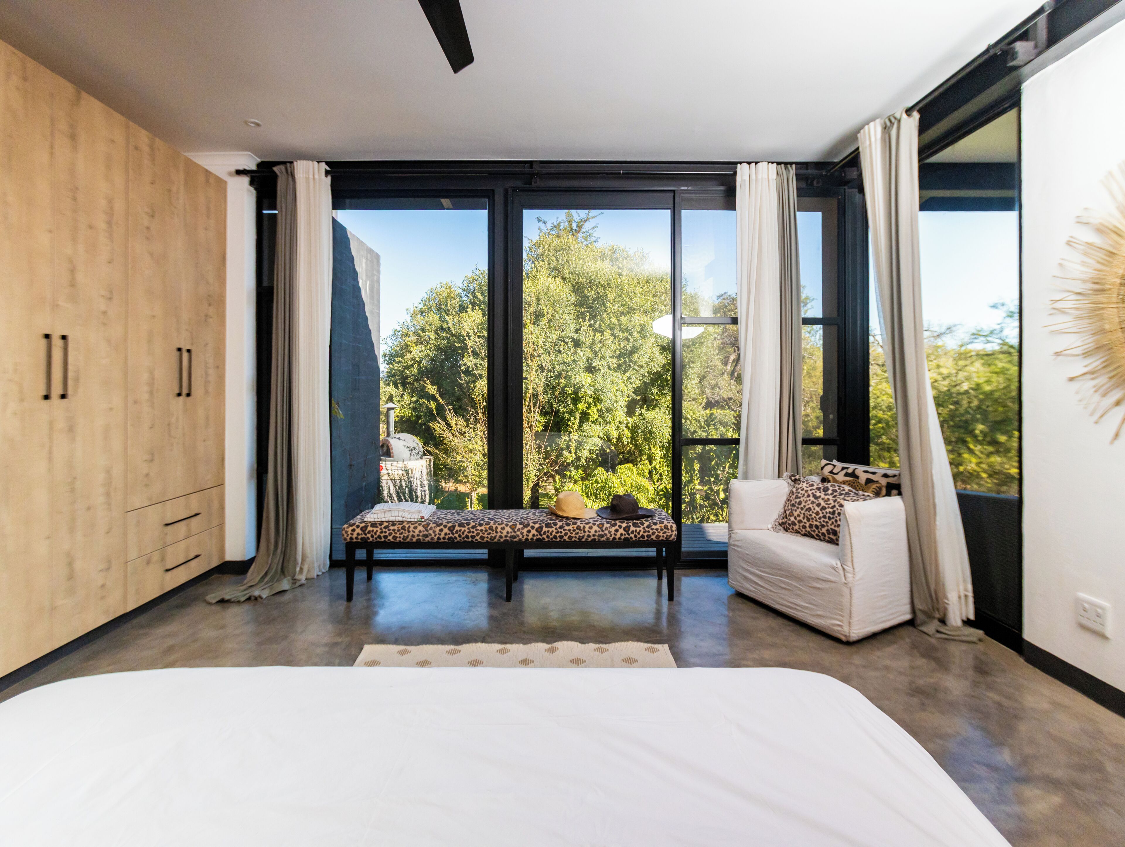 Quarto moderno com uma cama grande, um sofá e vista para o verde através de janelas do chão ao teto.