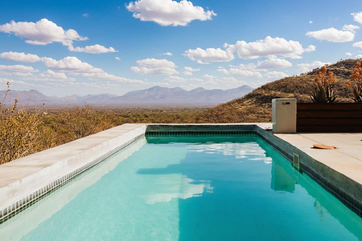 A swimming pool with clear blue water, surrounded by a concrete deck, set against a backdrop of mountains and a clear sky.