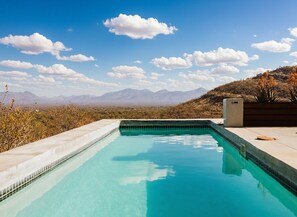 Une piscine à l’eau d’un bleu cristallin, entourée d’une terrasse en béton, avec en toile de fond des montagnes et un ciel dégagé.
