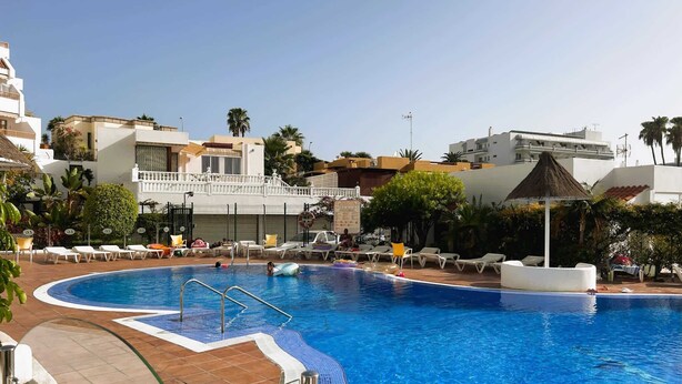 Une piscine avec une terrasse carrelée, des chaises longues et un bâtiment doté de balcons en arrière-plan.