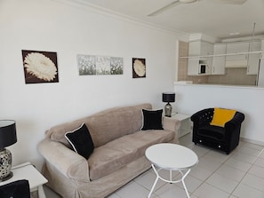 Ein Wohnzimmer mit einem beigen Sofa, einem kleinen weißen Tisch, einem schwarzen Sessel und Bildern an der Wand.