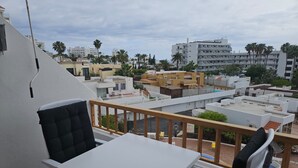 Un balcon avec une table blanche et une chaise noire, donnant sur un quartier résidentiel avec de nombreux immeubles et des palmiers.