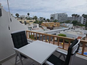Ein Balkon mit weißem Tisch und schwarzem Stuhl, mit Blick auf ein Wohngebiet mit mehreren Gebäuden und Palmen.