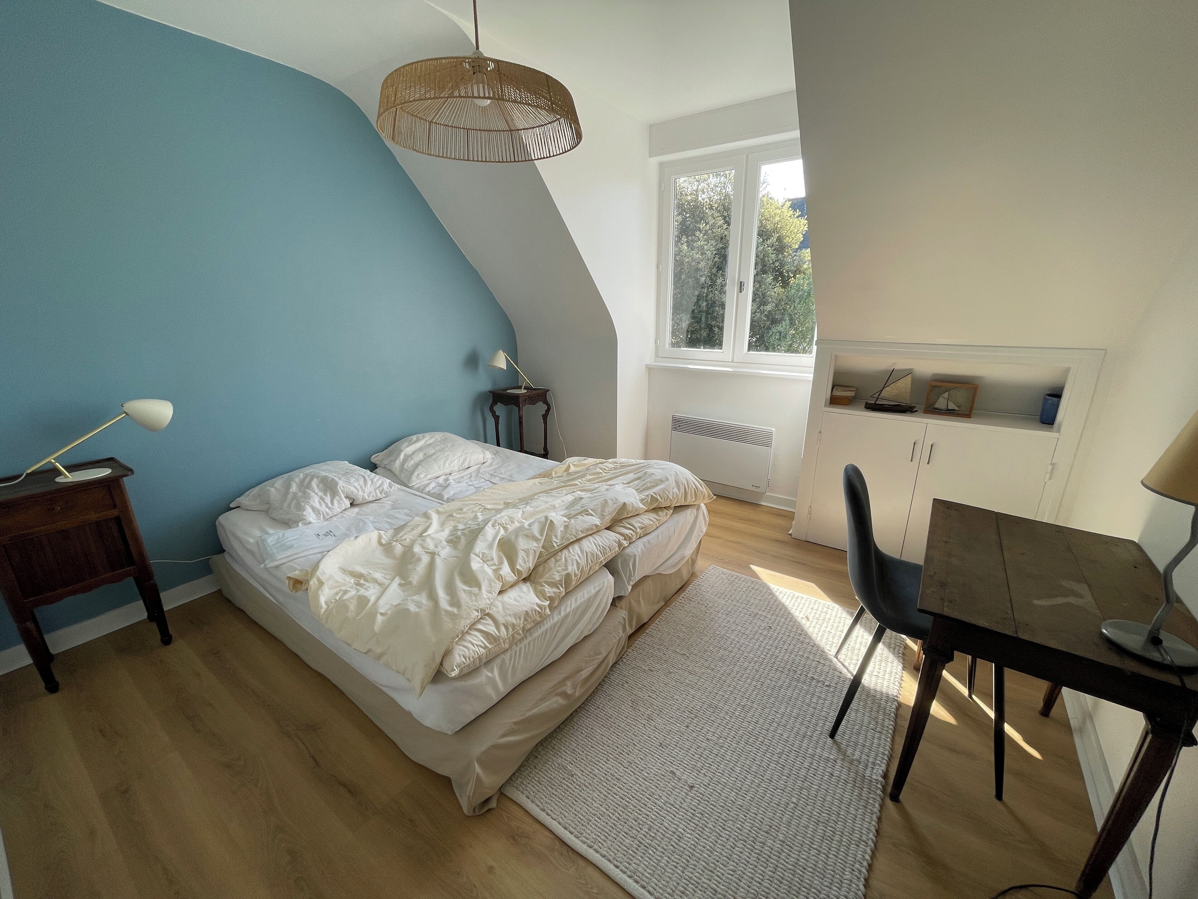 Een slaapkamer met een bed, nachtkastjes, een bureau, een stoel, een hanglamp en een raam.