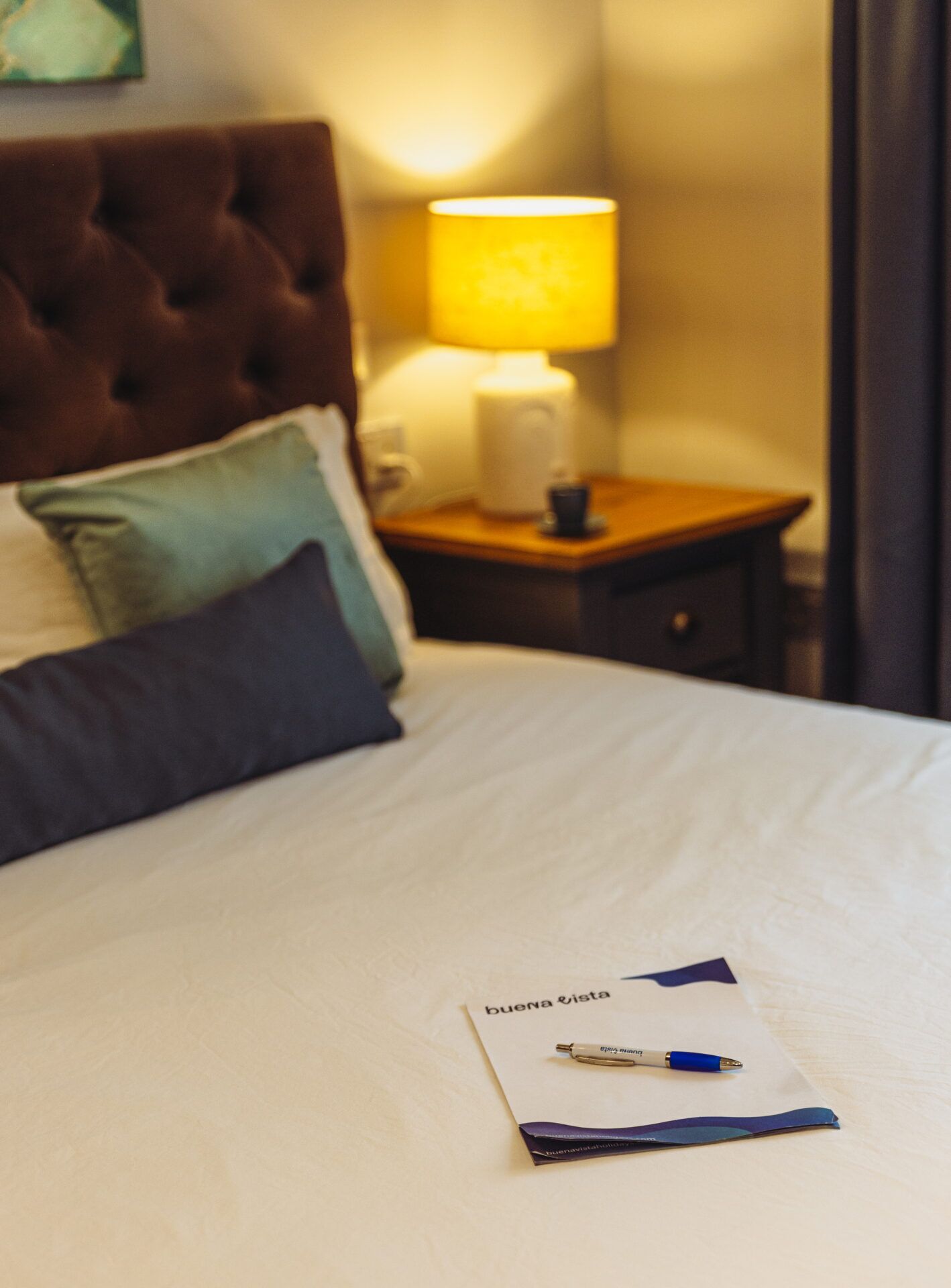 Une chambre d’hôtel avec un lit, des oreillers, une table de chevet avec une lampe et une carte accompagnée d’un stylo.