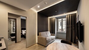 Una habitación de hotel moderna con piso de madera, una mesa redonda de mármol, un sofá color beige, un televisor montado en la pared y una ventana con cortinas.