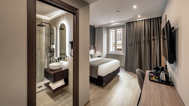 Una habitación de hotel moderna con una cama grande, un escritorio con hervidor y un baño con espejo grande y lavamanos.