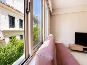Eine moderne Wohnung mit einem großen Fenster, einem rosafarbenen Sofa, einem Fernseher auf einem Holzboden und einem Balkon mit Grünpflanzen.