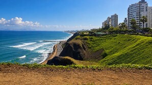 A coastal view with a beach, ocean waves, and a cityscape with high-rise buildings. - Spacious 3-Bedroom Apartment in Miraflores | Fast WiFi | Parking | Near Ocean View (Miraflores)