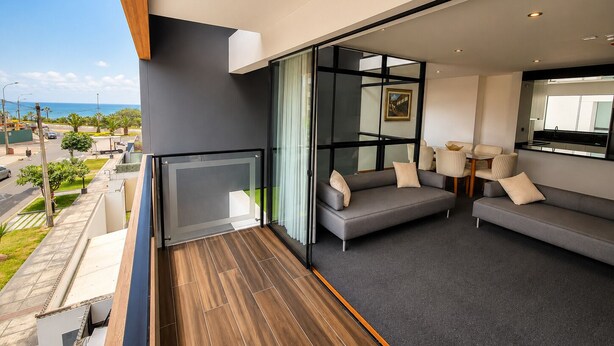A modern living room with a balcony, grey sofas, a dining area, and a view of the sea. - Spacious 3-Bedroom Apartment in Miraflores | Fast WiFi | Parking | Near Ocean View (Miraflores)