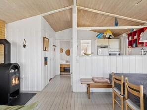 Ein gemütliches Interieur mit Kaminofen, einem Essbereich mit Bank und Stühlen, einer Küche mit Schränken und einem roten Regal sowie einem Schlafzimmer mit Bett und hängender Körbchen.