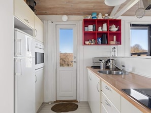 Eine Küche mit weißen Schränken, einer Holzausziehtafel, einem roten Regal mit Küchenutensilien und Blick durch die Tür.