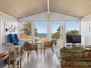 Ein gemütliches Zimmer mit Holzboden, großen Fenstern, einem Fernseher, Korbmöbeln und Blick auf das Meer.