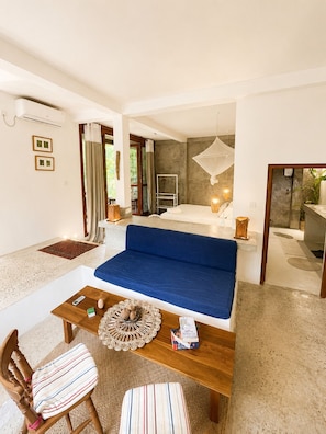 Una habitación con cama, sofá, mesa y silla. Hay una ventana con cortinas, una mesita de noche con lámpara y un aire acondicionado instalado en la pared.