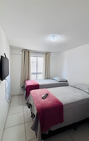 Une chambre avec deux lits, un téléviseur fixé au mur et une fenêtre avec des rideaux.
