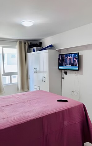 A room with a bed covered in pink, a television mounted on the wall, and a window with curtains. - Conforto em Cabo Branco 2 Qtos - Piscina (João Pessoa)