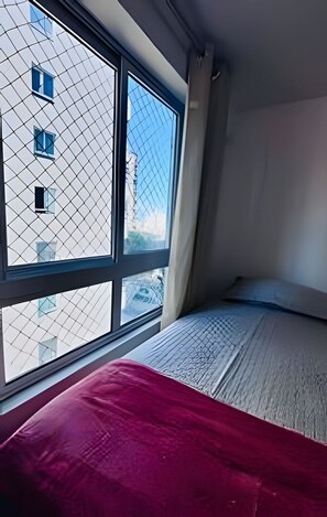 A bedroom with a large window, a bed with a red blanket, and a view of a building with multiple windows. - Conforto em Cabo Branco 2 Qtos - Piscina (João Pessoa)