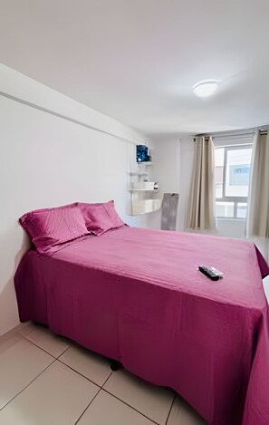 A single-bed room with a pink bedspread, a remote control on the bed, a window with curtains, and a white wall with a shelf. - Conforto em Cabo Branco 2 Qtos - Piscina (João Pessoa)