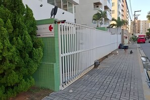 A gated entrance with a green fence, a paved sidewalk, and a red vehicle parked on the street. - Conforto em Cabo Branco 2 Qtos - Piscina (João Pessoa)