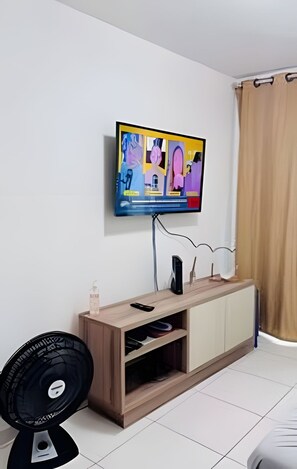 A modern living room with a flat-screen TV mounted on the wall, a wooden entertainment unit below, and a black fan on the floor. - Conforto em Cabo Branco 2 Qtos - Piscina (João Pessoa)