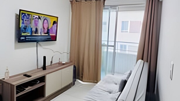 A modern bedroom with a bed, a TV mounted on the wall, and a window with curtains. - Conforto em Cabo Branco 2 Qtos - Piscina (João Pessoa)