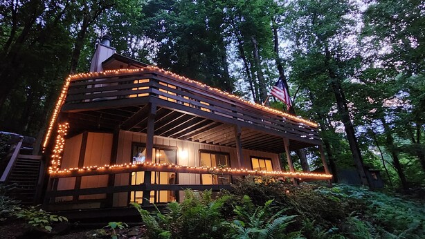 Une cabane en bois dotée d’un porche, entourée d’arbres et illuminée de guirlandes lumineuses.